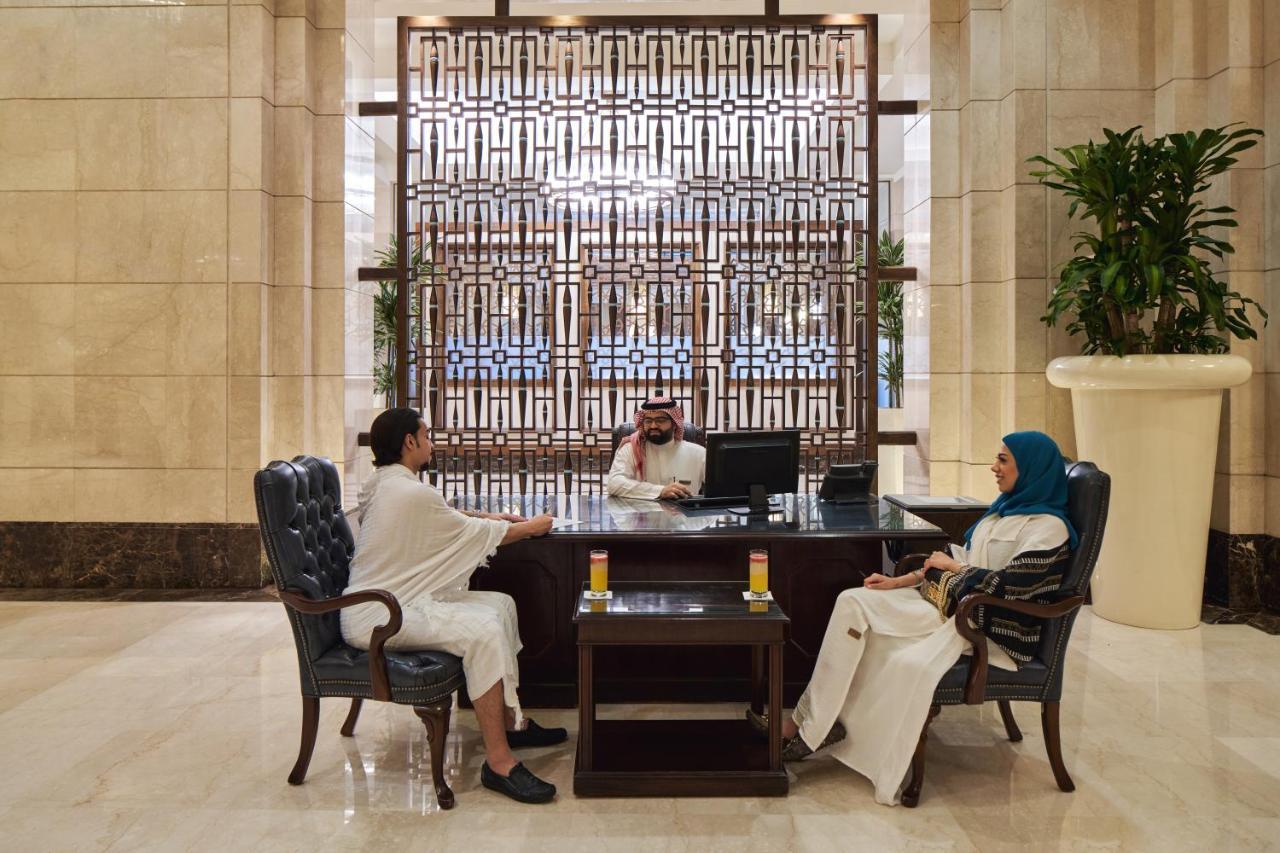 Makkah Clock Royal Tower, A Fairmont Hotel La La Mecca Esterno foto The photo shows a reception area in a hotel or a similar establishment. In the foreground, there are two individuals sitting in upholstered chairs. One person is wearing traditional attire, while the other is dressed in a modest outfit with a headsca
