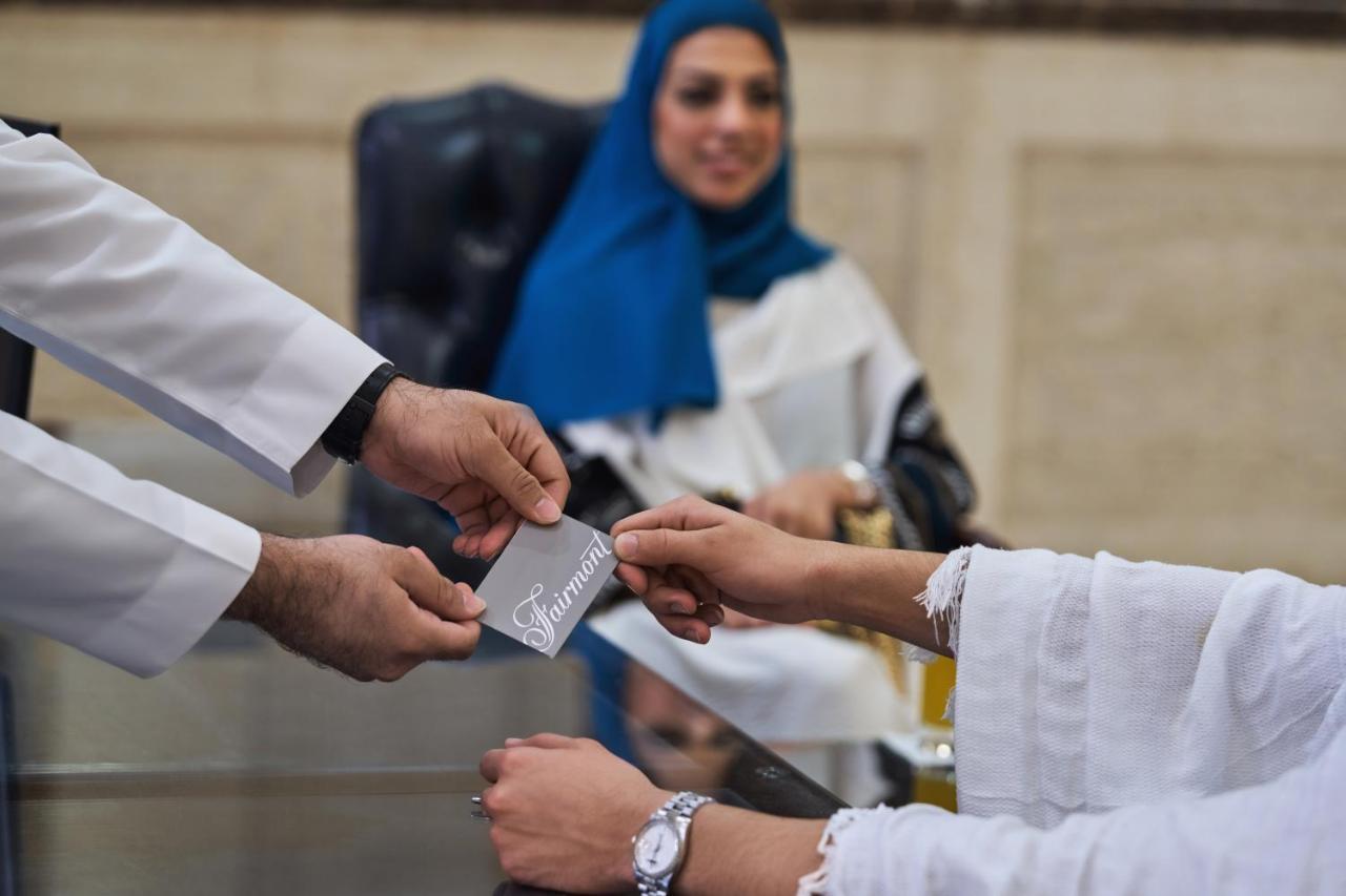 Makkah Clock Royal Tower, A Fairmont Hotel La La Mecca Esterno foto The photo shows a professional setting where a person in a white coat is handing a card or document to another person. The recipient, who is wearing a traditional outfit and a watch, appears to be engaged in a conversation or transaction. In the back