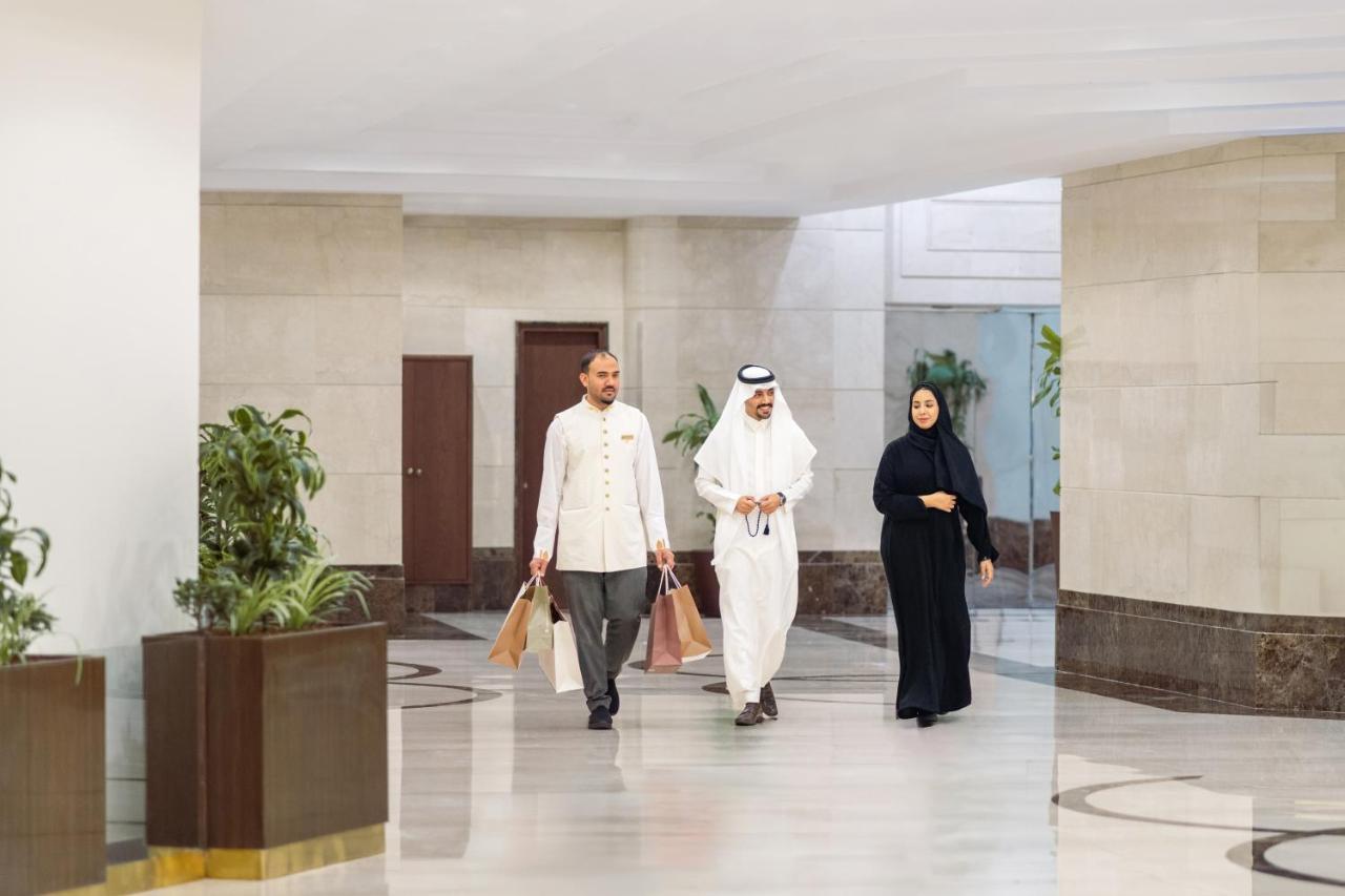 Makkah Clock Royal Tower, A Fairmont Hotel La La Mecca Esterno foto The photo shows three individuals walking through a modern indoor space. On the left, there is a man wearing a white traditional outfit with dark pants and carrying bags. In the center, another man is dressed in a long white thobe and a headscarf. To