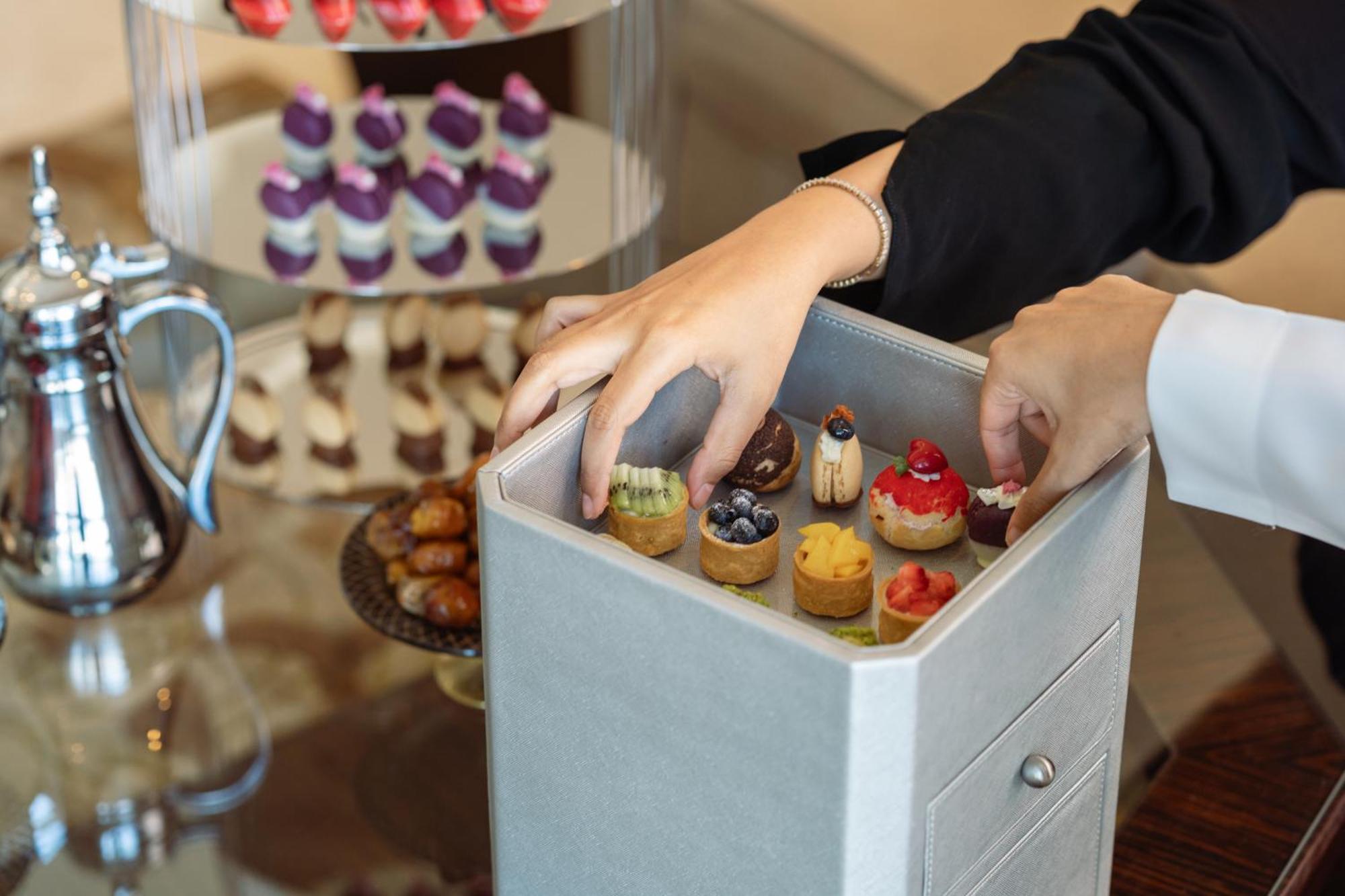 Makkah Clock Royal Tower, A Fairmont Hotel La La Mecca Esterno foto The photo depicts a person’s hands reaching into a gray box filled with an assortment of small, intricate pastries or desserts. These treats appear to be delicately crafted, showcasing various toppings and colors, suggesting they are meant for a spec