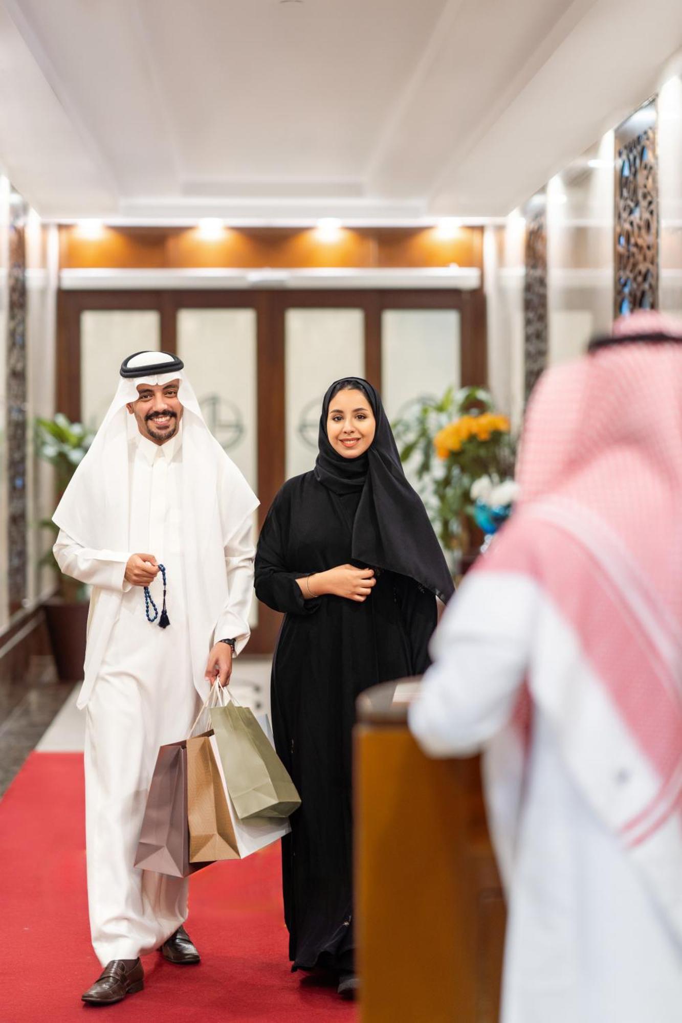 Makkah Clock Royal Tower, A Fairmont Hotel La La Mecca Esterno foto The photo shows a man and a woman entering a building. The man is wearing a traditional white thobe and a black agal, while the woman is dressed in a black abaya. Both are smiling and appear to be in a welcoming and pleasant environment. The backgrou