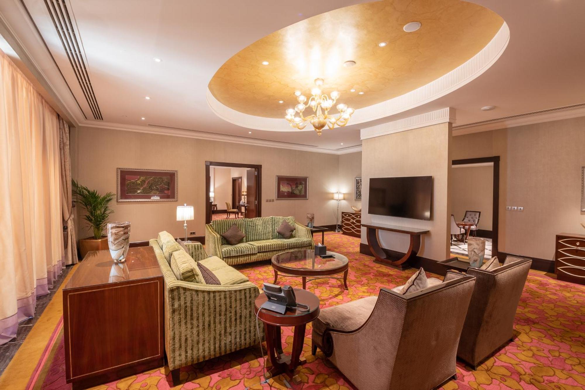 Makkah Clock Royal Tower, A Fairmont Hotel La La Mecca Esterno foto The photo shows a spacious and elegantly designed living room. The room features a light-colored ceiling with a decorative circular design and a chandelier. There are plush sofas in a green fabric arranged around a central coffee table, along with tw