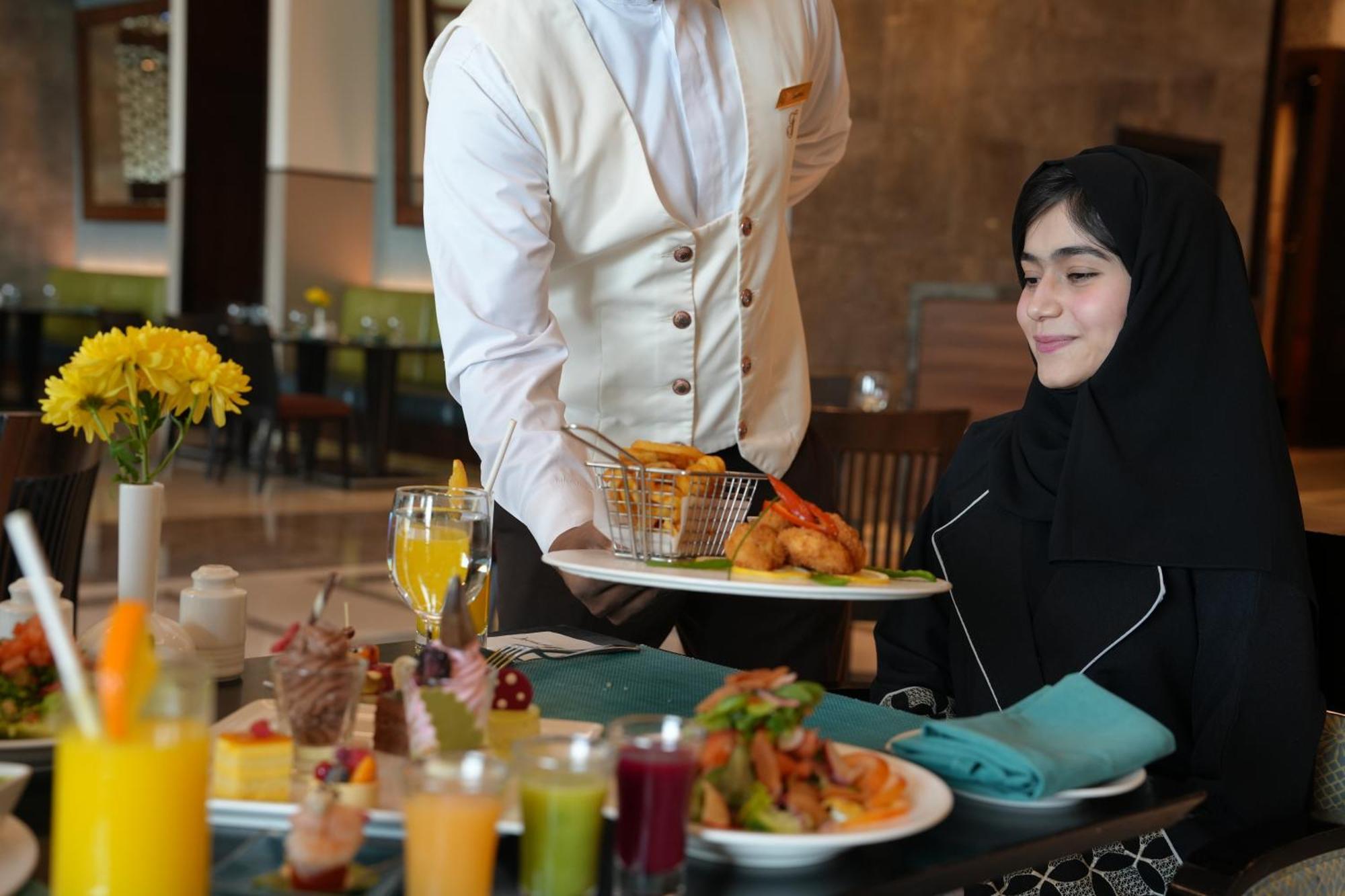 Makkah Clock Royal Tower, A Fairmont Hotel La La Mecca Esterno foto The image shows a dining setting in a restaurant. A waiter, dressed in a formal outfit, is serving food to a woman wearing a black hijab. The table is filled with a variety of colorful dishes and drinks, including fruit juices and desserts. There are