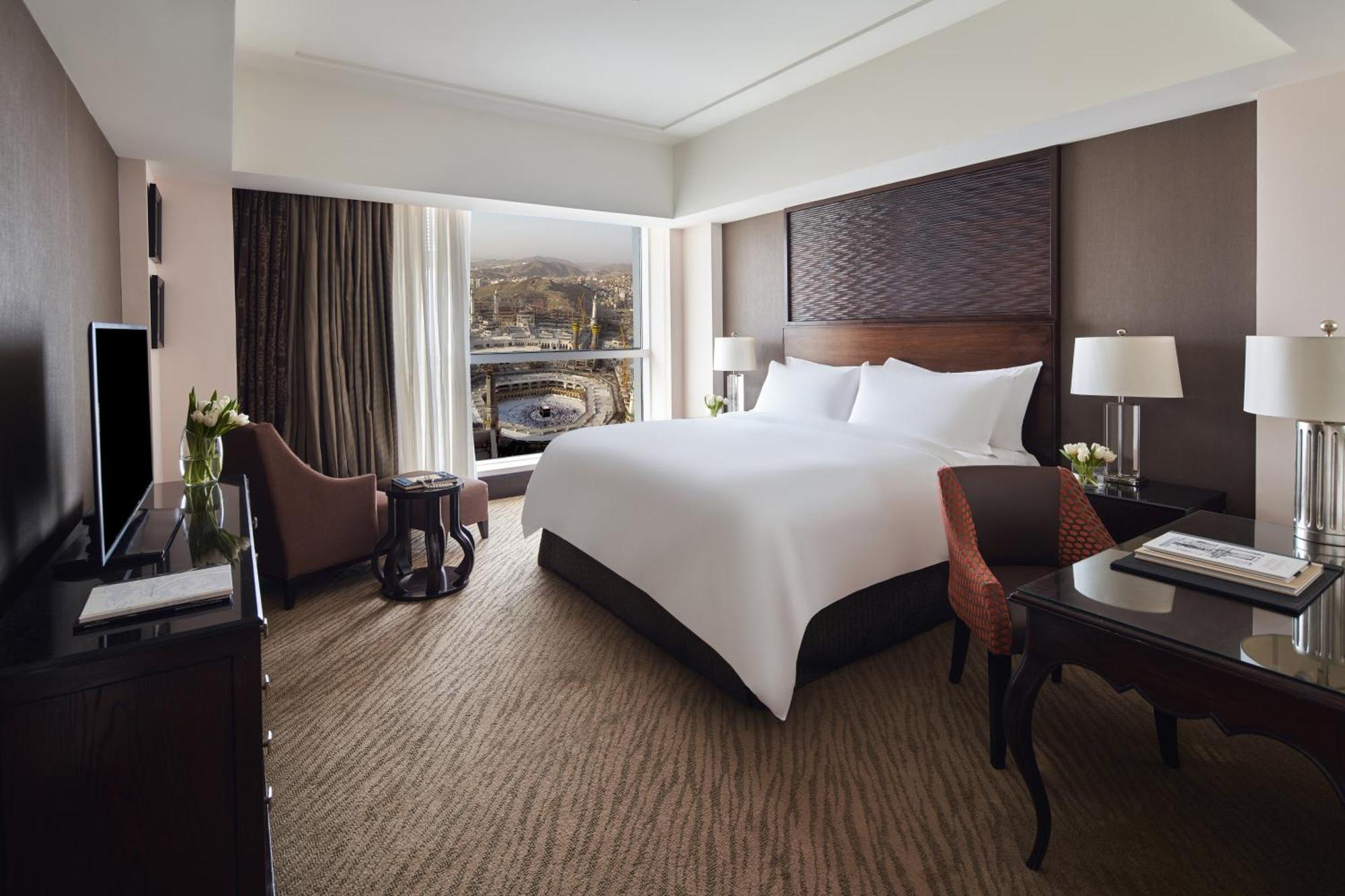 Makkah Clock Royal Tower, A Fairmont Hotel La La Mecca Esterno foto The photo shows a modern hotel room featuring a large bed with white linens, a stylish headboard, and two bedside lamps. There's a cozy seating area with two chairs, one of which is upholstered in orange fabric. A small table is present near the chai