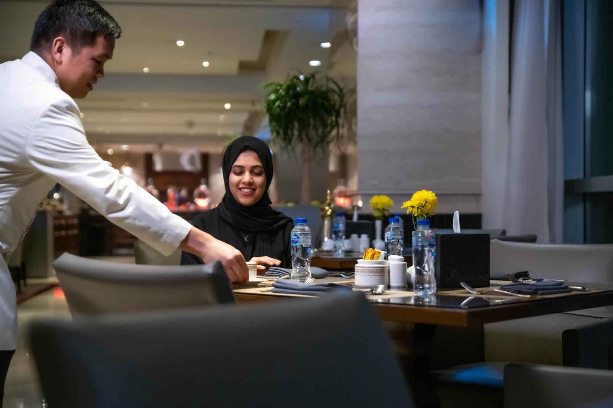 Makkah Clock Royal Tower, A Fairmont Hotel La La Mecca Esterno foto The photo shows a restaurant setting where a waiter is serving a woman sitting at a dining table. The woman is wearing a black outfit and a hijab, and she is smiling as the waiter interacts with her. The table is set with water bottles, a flower arra