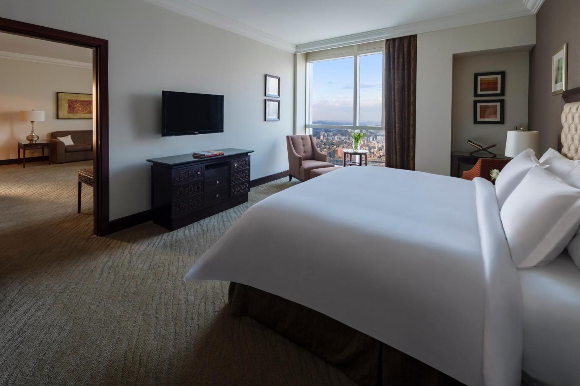 Makkah Clock Royal Tower, A Fairmont Hotel La La Mecca Esterno foto The photo shows a modern hotel room featuring a large bed with white linens positioned prominently in the foreground. To the left, there is a dark wooden dresser with a flat-screen TV mounted on the wall above it. On the right side of the room, a lar