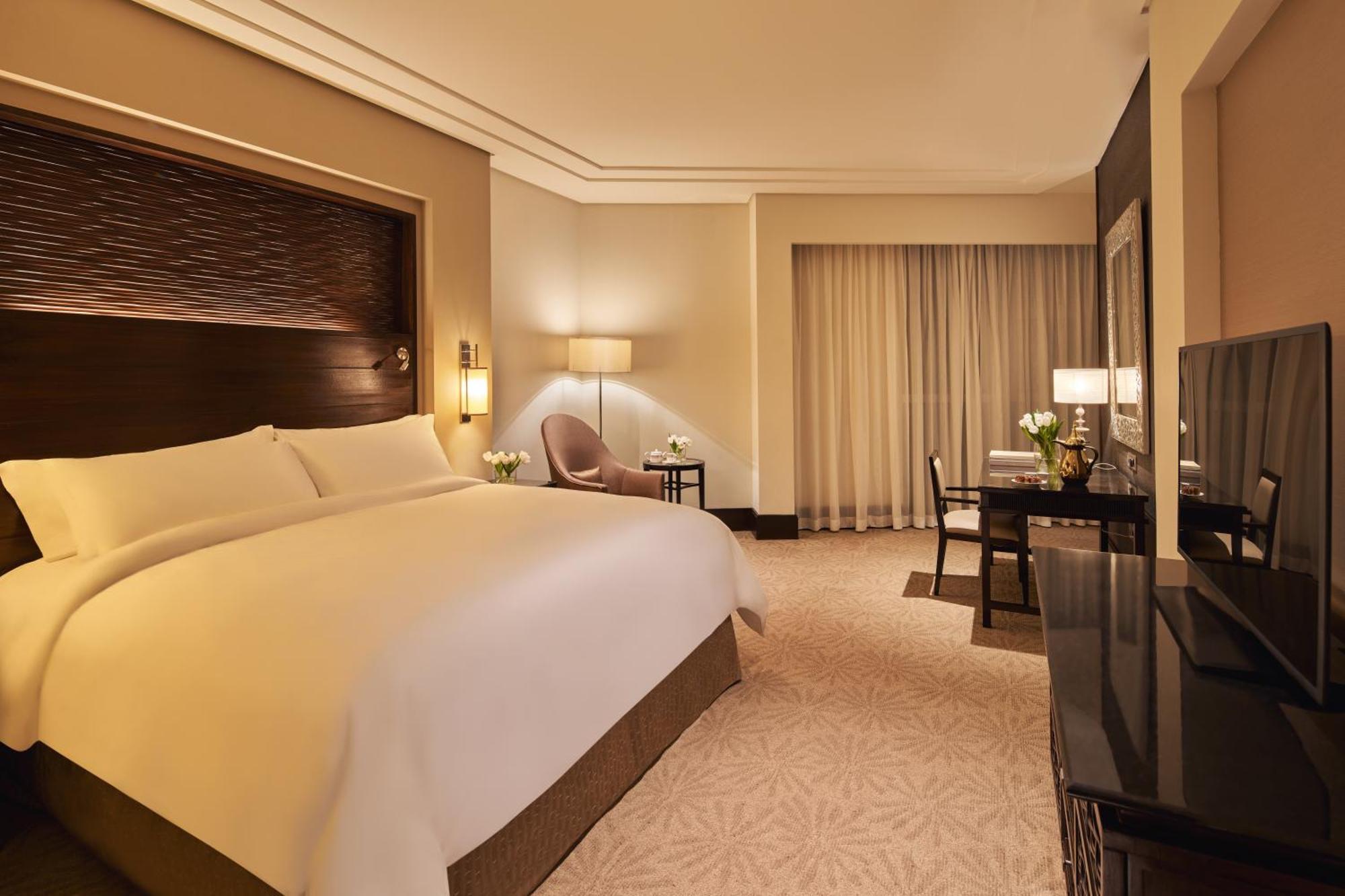 Makkah Clock Royal Tower, A Fairmont Hotel La La Mecca Esterno foto The photo shows a hotel room featuring a spacious and elegantly designed interior. In the foreground, there is a large bed with white bedding and a decorative headboard. To the side, there is a small table and a chair, creating a cozy seating area. A