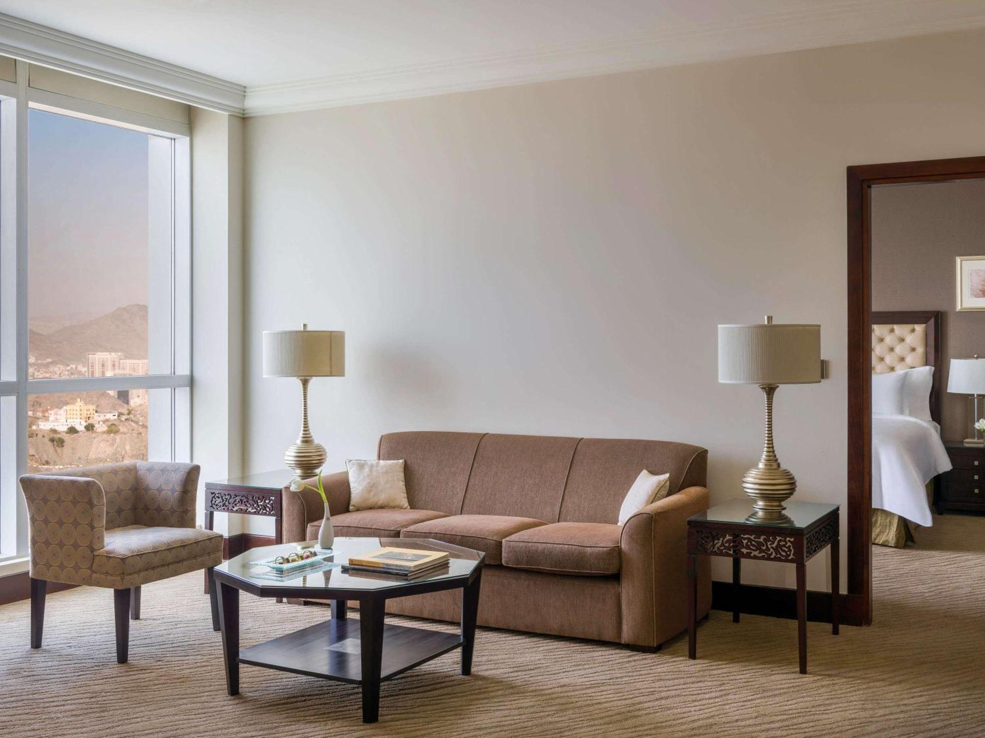 Makkah Clock Royal Tower, A Fairmont Hotel La La Mecca Esterno foto The photo shows a modern hotel suite living area. It features a brown sofa, a small glass-topped coffee table, and a patterned armchair. There are two stylish lamps on either side of the sofa. A large window lets in natural light, offering a view of 