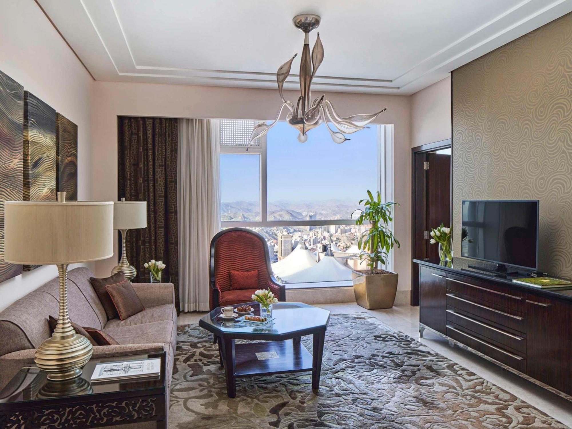 Makkah Clock Royal Tower, A Fairmont Hotel La La Mecca Esterno foto The photo shows a luxurious living room with a modern design. It features a large window that offers a view of the cityscape. The room is furnished with a comfortable sofa, an accent chair, and a coffee table placed on an ornate rug. A stylish chande