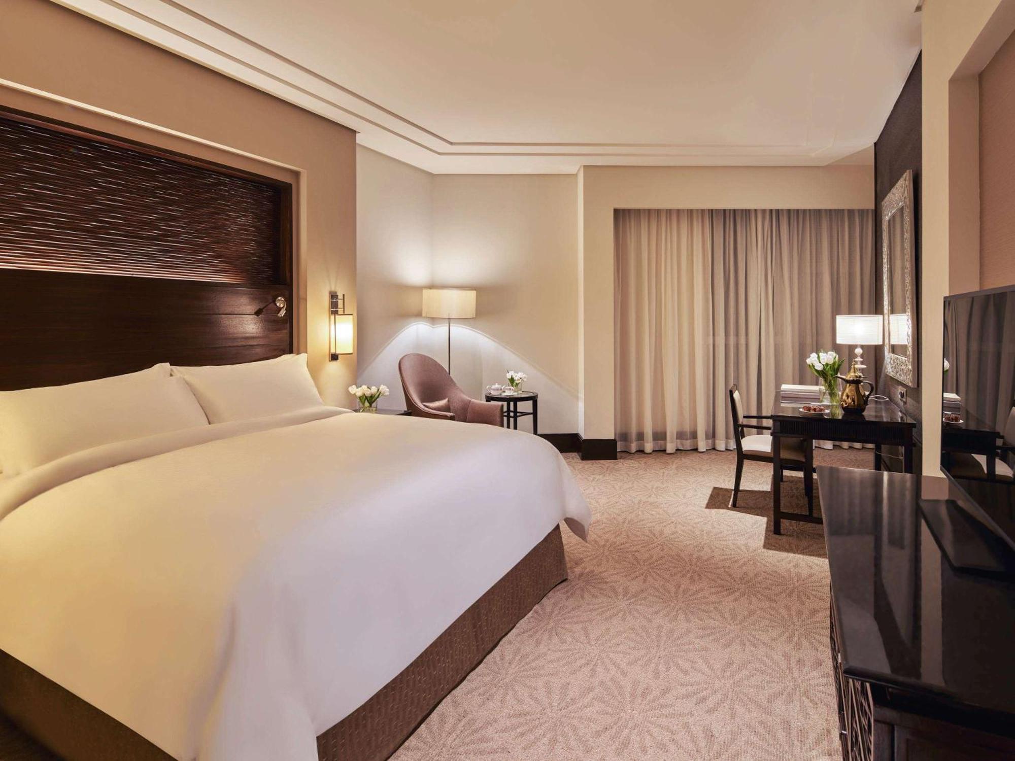 Makkah Clock Royal Tower, A Fairmont Hotel La La Mecca Esterno foto The photo shows a spacious hotel room featuring a large bed with white linens and a stylish headboard. To the side of the bed, there is a bedside lamp and a comfortable armchair. A window with sheer curtains allows natural light to enter the room. In