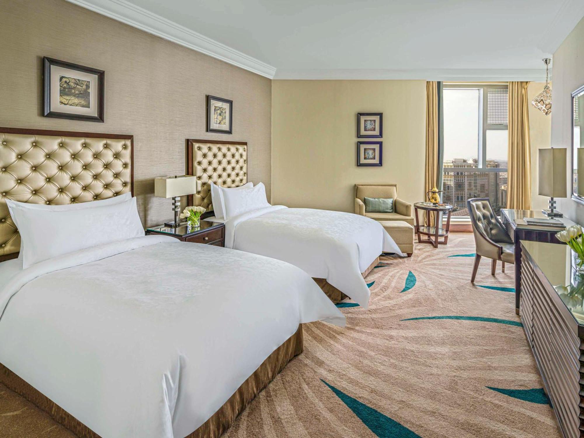 Makkah Clock Royal Tower, A Fairmont Hotel La La Mecca Esterno foto The photo shows a stylish hotel room featuring two beds with tufted headboards. The beds are dressed in white linens, giving a fresh and clean appearance. Next to each bed, there are bedside tables with lamps. There is a small seating area with a cha