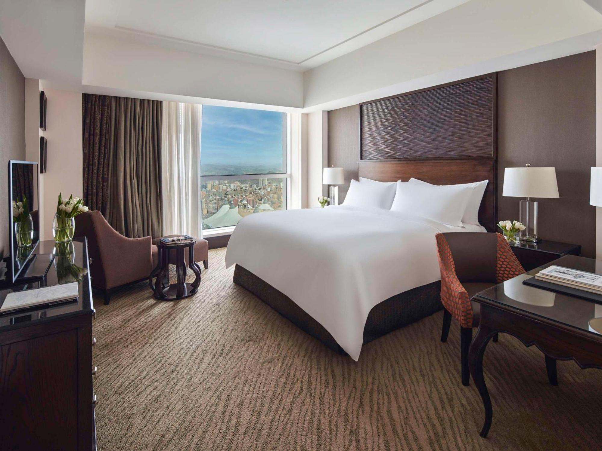Makkah Clock Royal Tower, A Fairmont Hotel La La Mecca Esterno foto The photo shows a hotel room featuring a large bed with white linens positioned against a wooden headboard. There is a large window providing natural light and a view outside. The room includes two bedside tables with lamps, a comfortable armchair, a