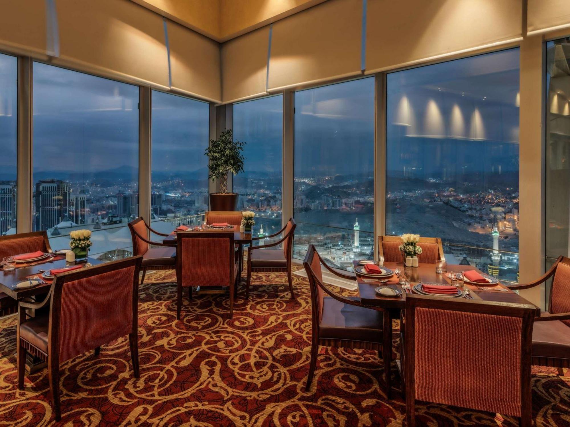 Makkah Clock Royal Tower, A Fairmont Hotel La La Mecca Esterno foto The photo shows an elegant restaurant interior with several tables and chairs arranged for dining. The restaurant features large windows offering a panoramic view of the city at dusk, showcasing a skyline illuminated by city lights. The decor include