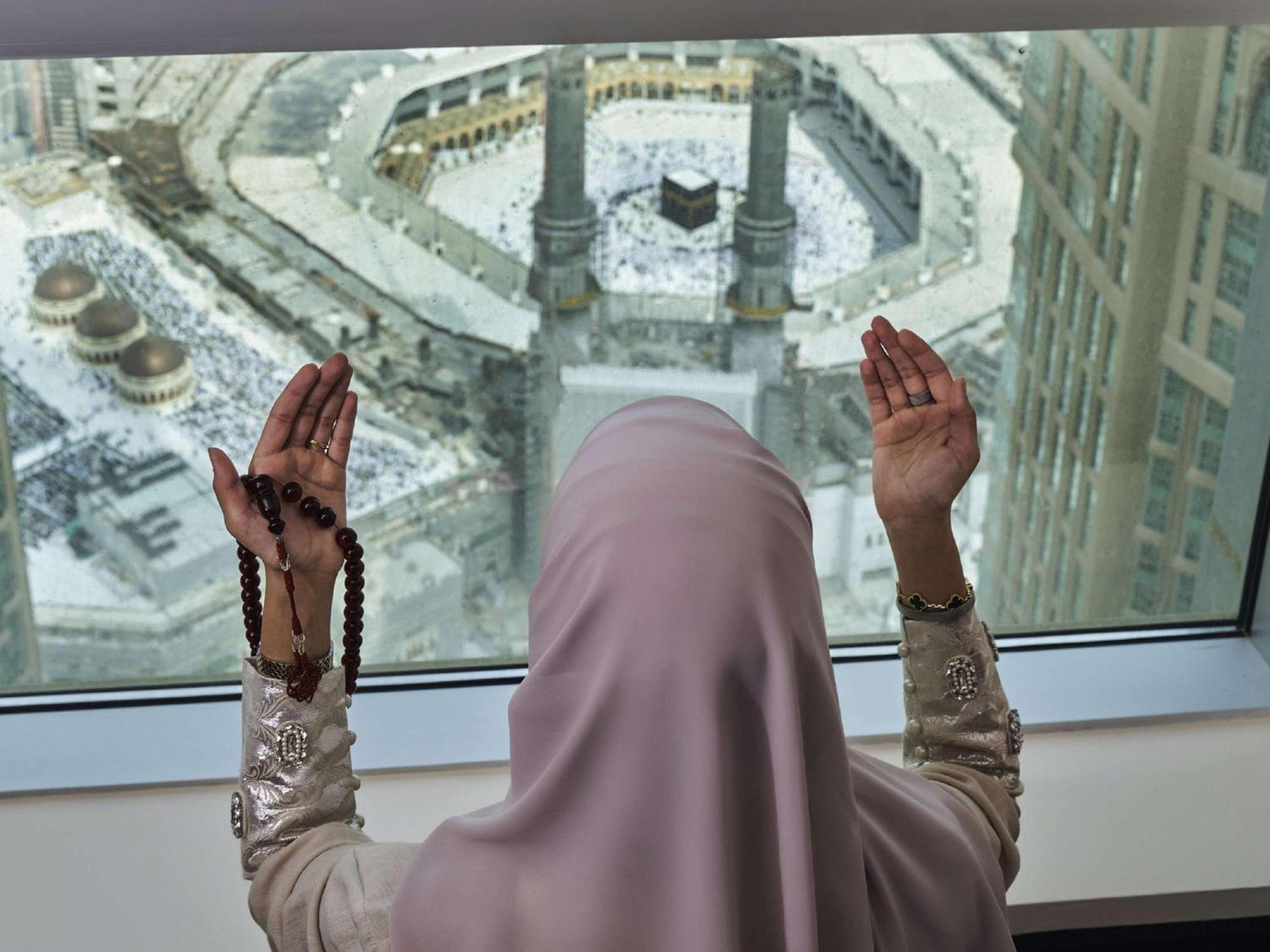 Makkah Clock Royal Tower, A Fairmont Hotel La La Mecca Esterno foto The photo shows a person wearing a hijab, positioned in front of a large window. They appear to be praying or in a moment of reflection, with their hands raised and holding prayer beads. Outside the window, the view reveals the Kaaba, an important st
